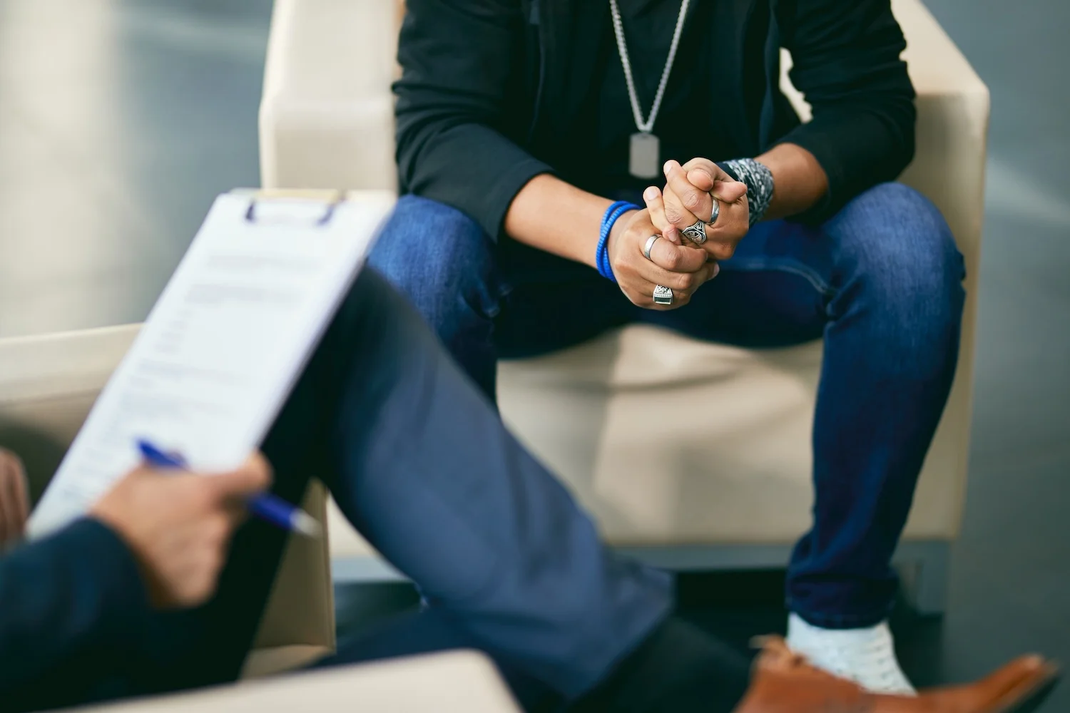 Close-up of a man sitting with clasped hands during a counseling session, speaking with a therapist holding a clipboard.
