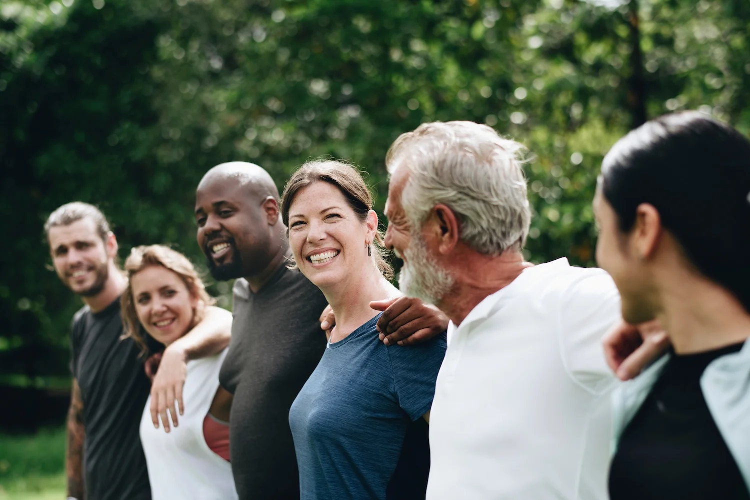 A diverse group of adults stands arm in arm outside, smiling and supporting each other.